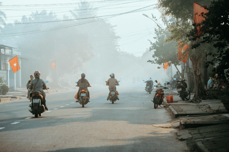 Three people riding scooters on a smoggy street.